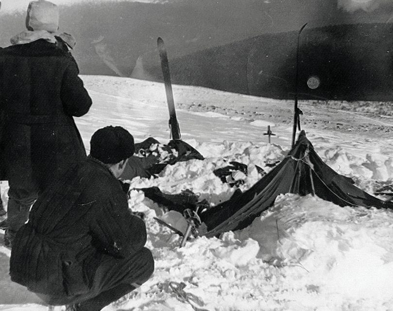 Black and white photo from February 1959: two searchers in heavy coats look at the Dyatlov group's tent, collapsed and half covered by snow on an open slope, with a ski standing upright in the snow