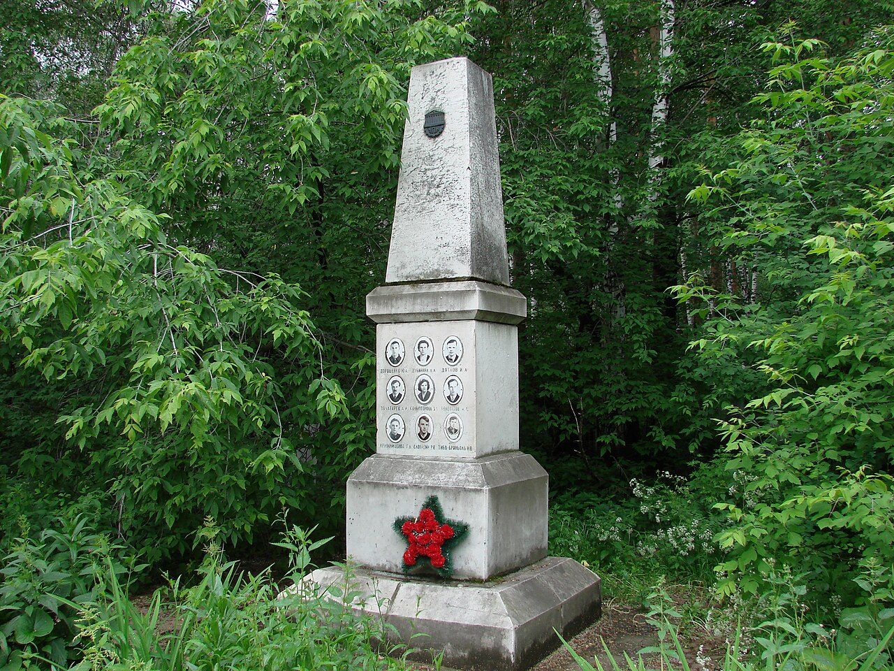 A grey stone obelisk among green trees at Mikhailovskoe Cemetery in Yekaterinburg, the memorial over the Dyatlov group's grave, with nine small portraits on its base and a red star wreath at the bottom
